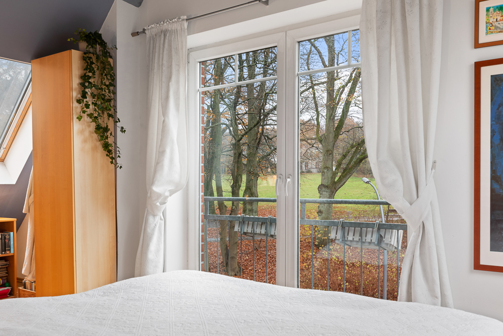 Helles Schlafzimmer mit großen Fenstern, weißen Vorhängen und Blick auf Bäume im Garten.