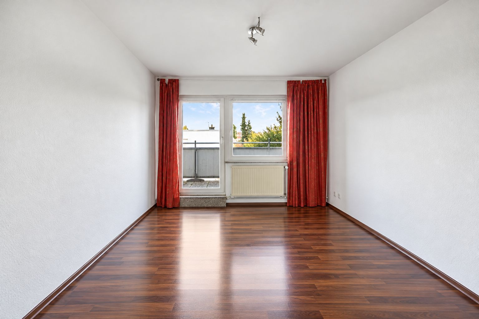 Leeres Zimmer mit Holzboden, weißen Wänden und Fenster mit roten Vorhängen zum Balkon.