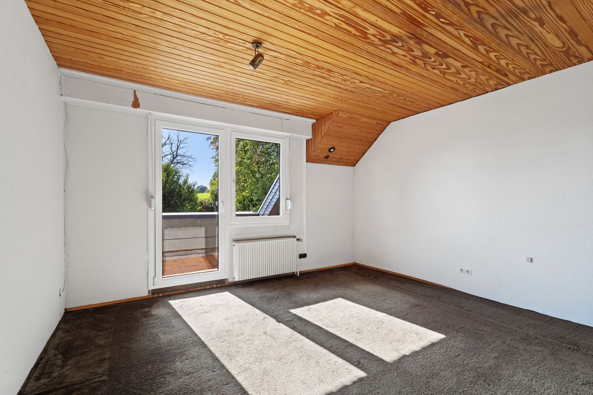 Leeres Zimmer mit Teppichboden, Holzdecke und Zugang zu einem kleinen Balkon durch eine Glastür.