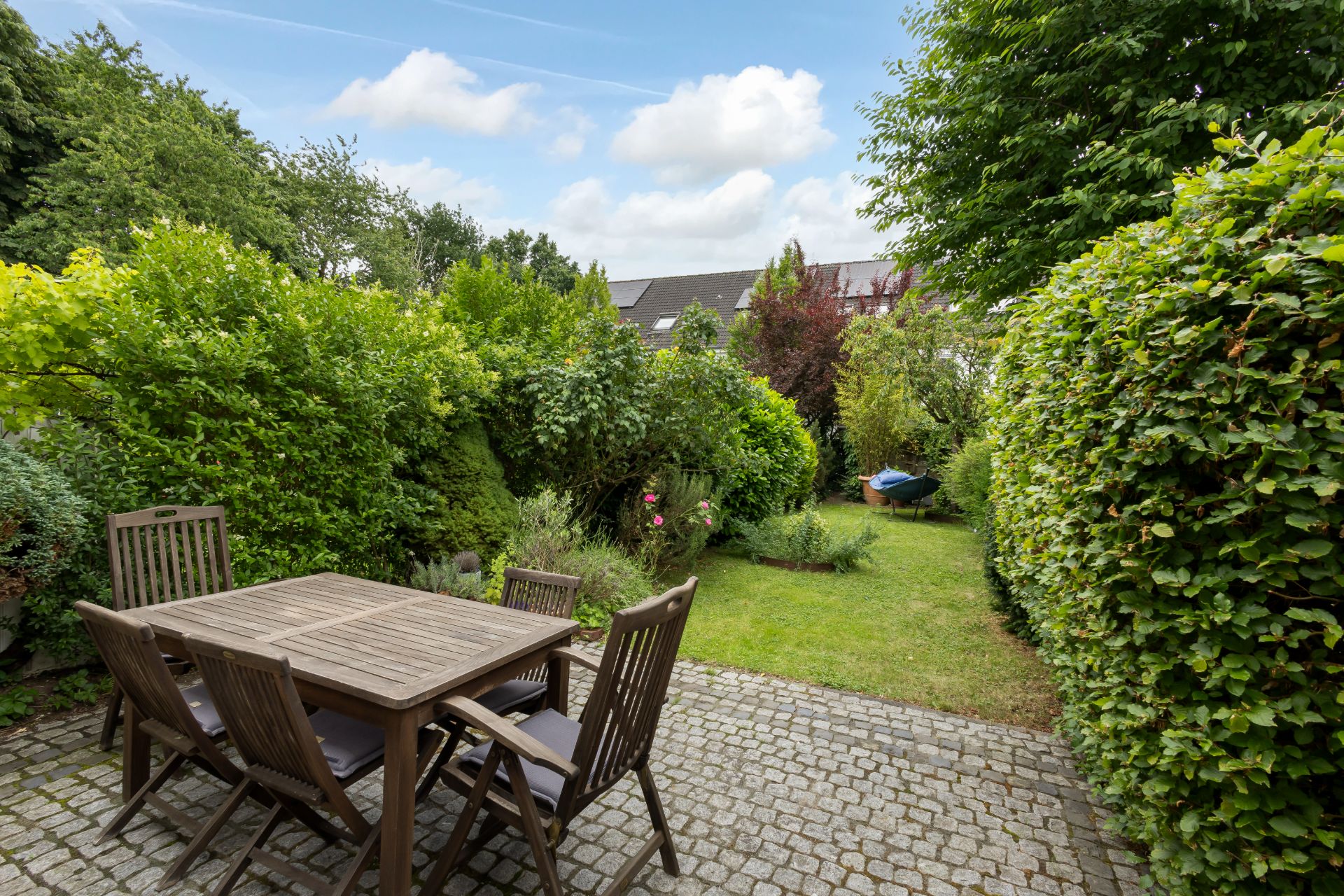 Gepflasterte Terrasse mit Holztisch und Stühlen, umgeben von grünem Garten und Büschen.
