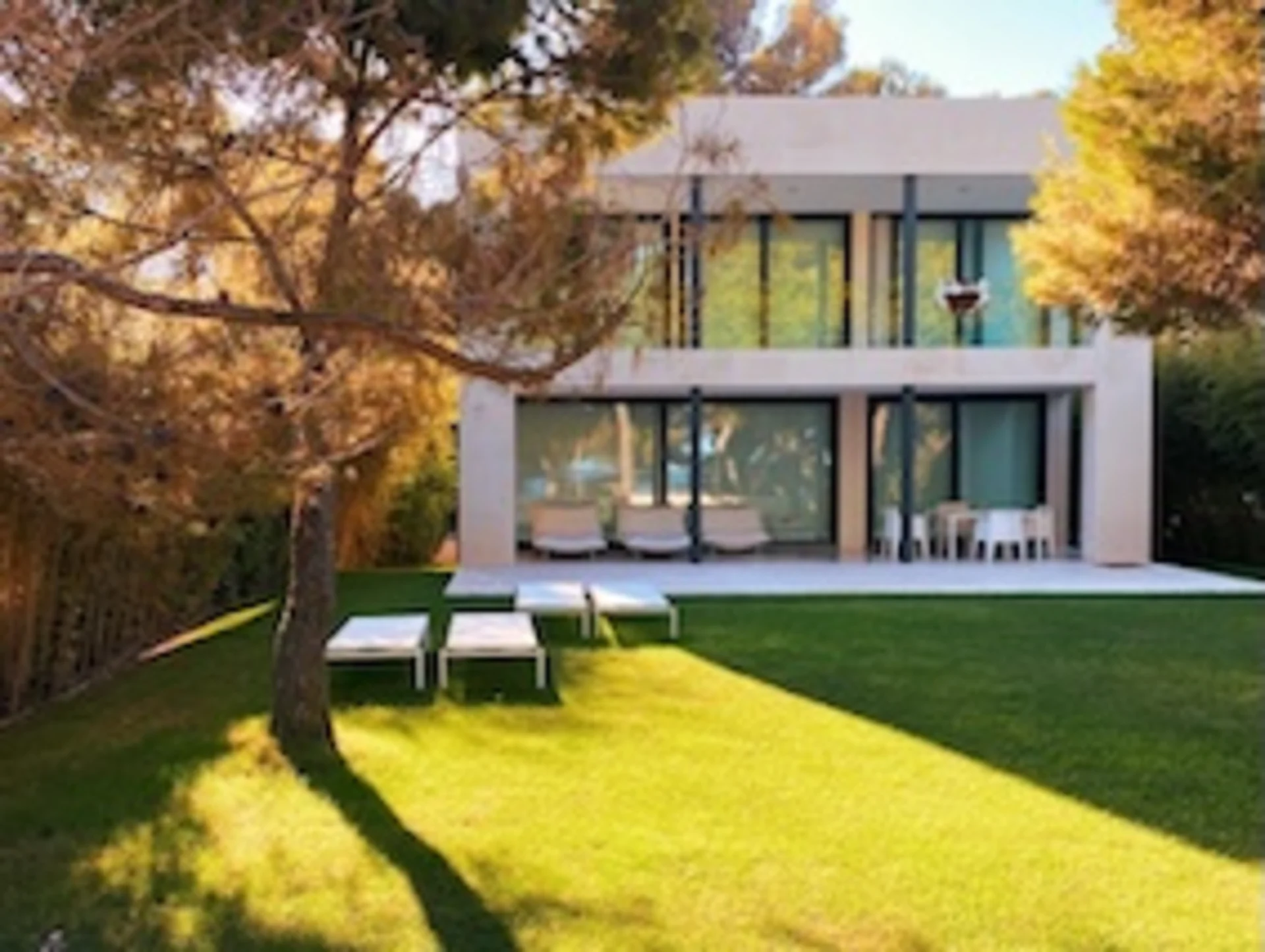 Moderne zweistöckige Wohnhausfassade mit großen Fenstern und Garten mit Liegen und Bäumen.