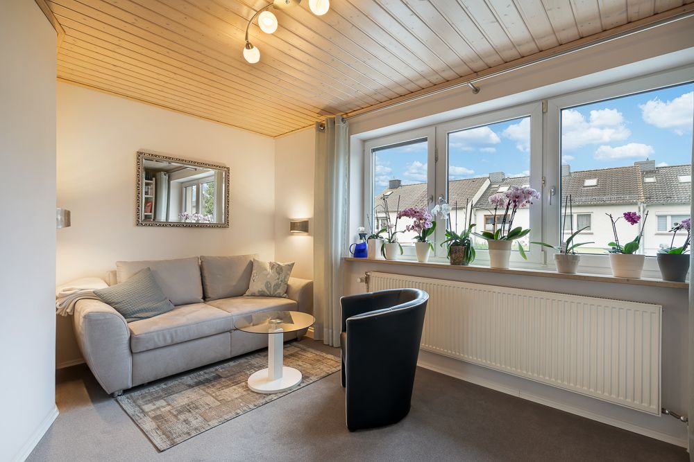 Helles Wohnzimmer mit Sofa, Sessel, kleinem Tisch und vielen Pflanzen auf der Fensterbank.