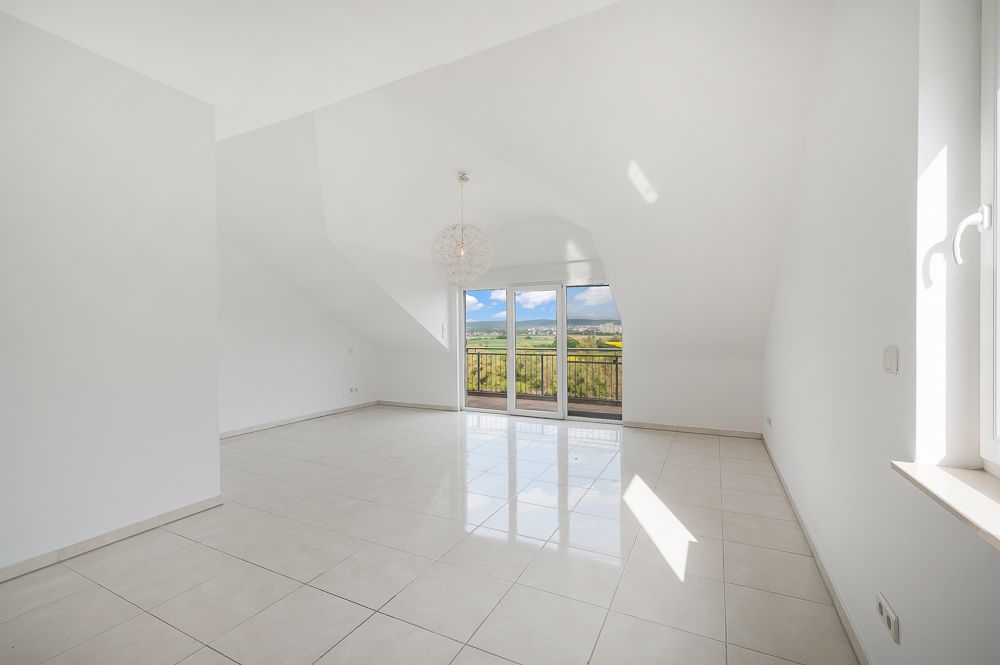 Heller, leerer Raum mit weißen Wänden, Fliesenboden und großer Fensterfront mit Blick ins Grüne.
