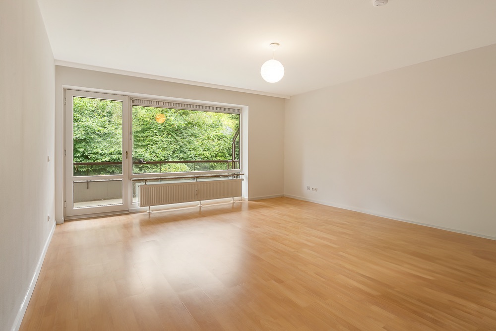Helles, leeres Wohnzimmer mit Holzfußboden, großem Fenster und Blick auf grüne Bäume.