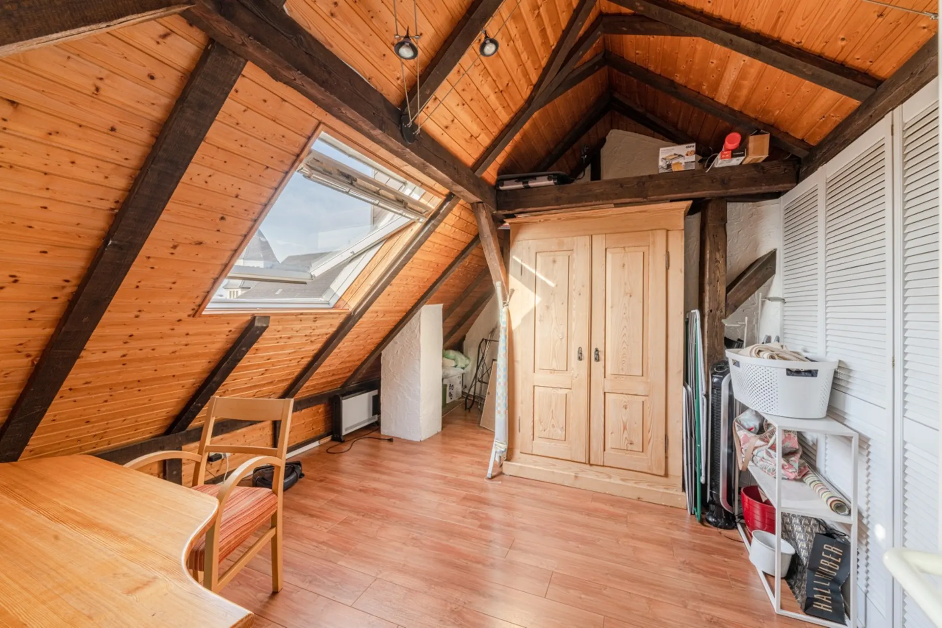 Helles Dachzimmer mit Holzdecke, Schrank, Regal und großem Fenster.