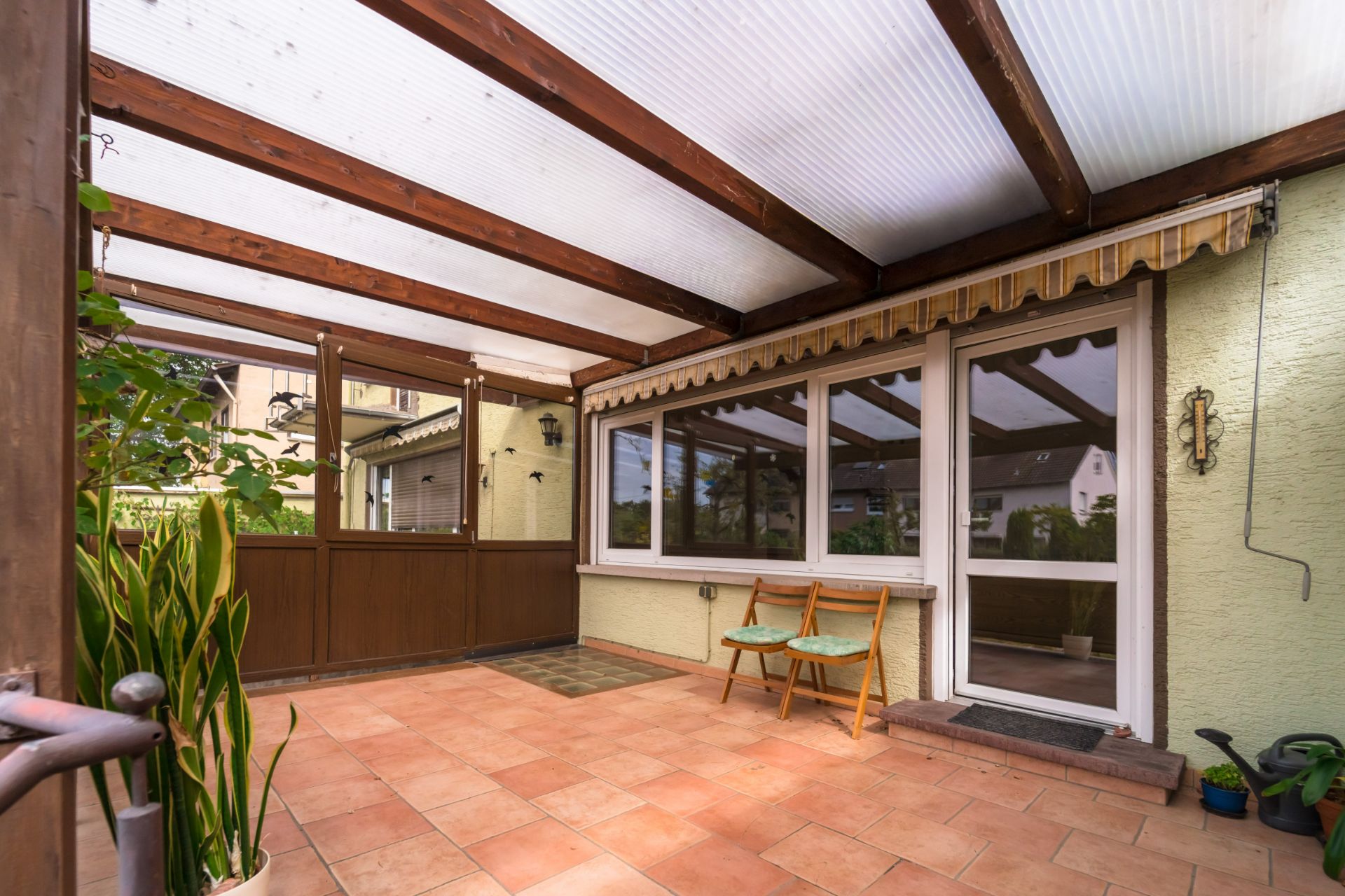 Überdachte Terrasse mit Fliesenboden, zwei Stühlen und großen Fenstern, Pflanzen an der Seite.