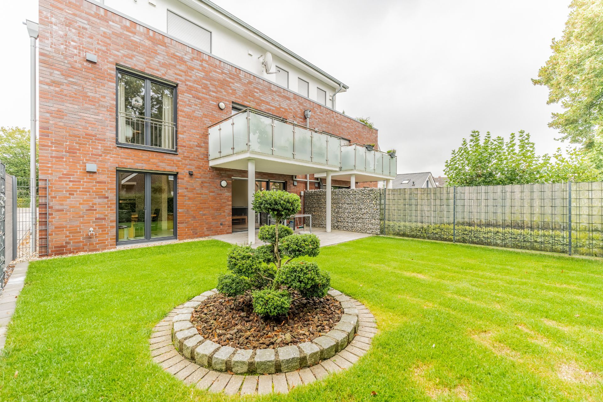 Moderner Garten mit Rasen, rundem Buschbeet und Backsteinhaus im Hintergrund.