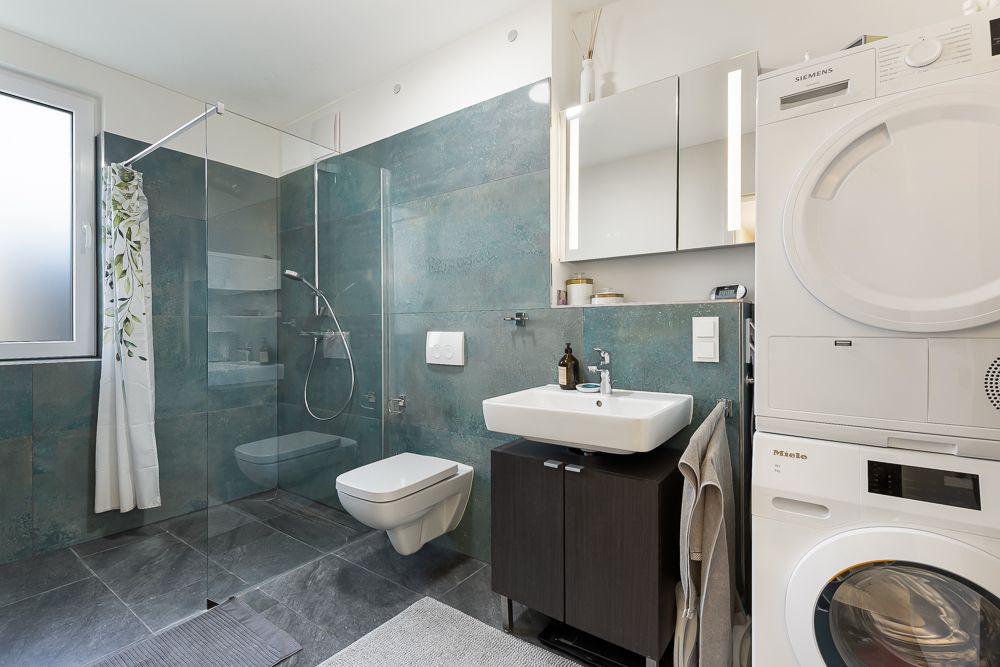 Modernes Badezimmer mit Dusche, WC, Waschbecken, Spiegel und Waschmaschine mit Trockner.