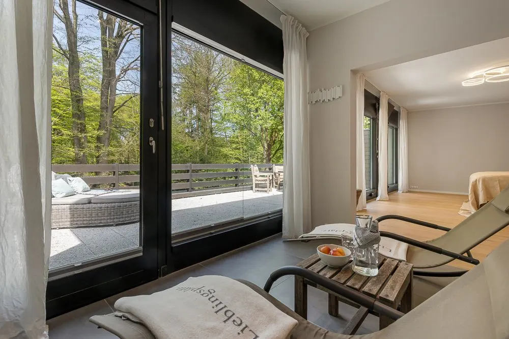 Helles Wohnzimmer mit Liegestühlen, Tisch, großer Glasfront und Blick auf Terrasse mit Sitzmöbeln.