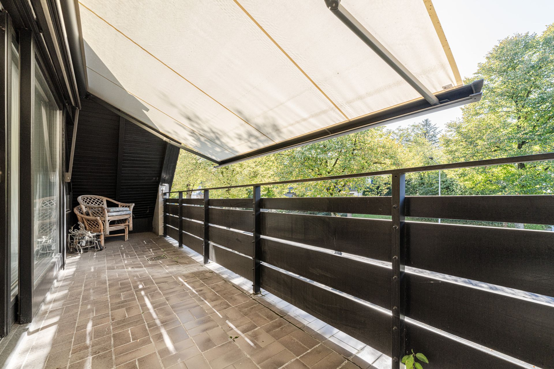 Überdachter Balkon mit dunklem Geländer, zwei Stühlen und Blick auf grüne Bäume.