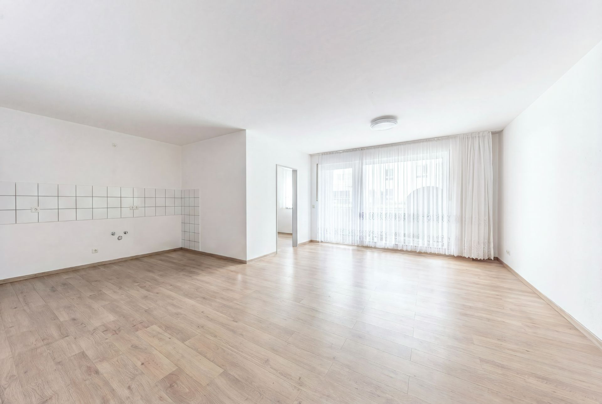 Heller, leerer Raum mit Parkettboden, großen Fenstern und weißem Fliesenspiegel an einer Wand.
