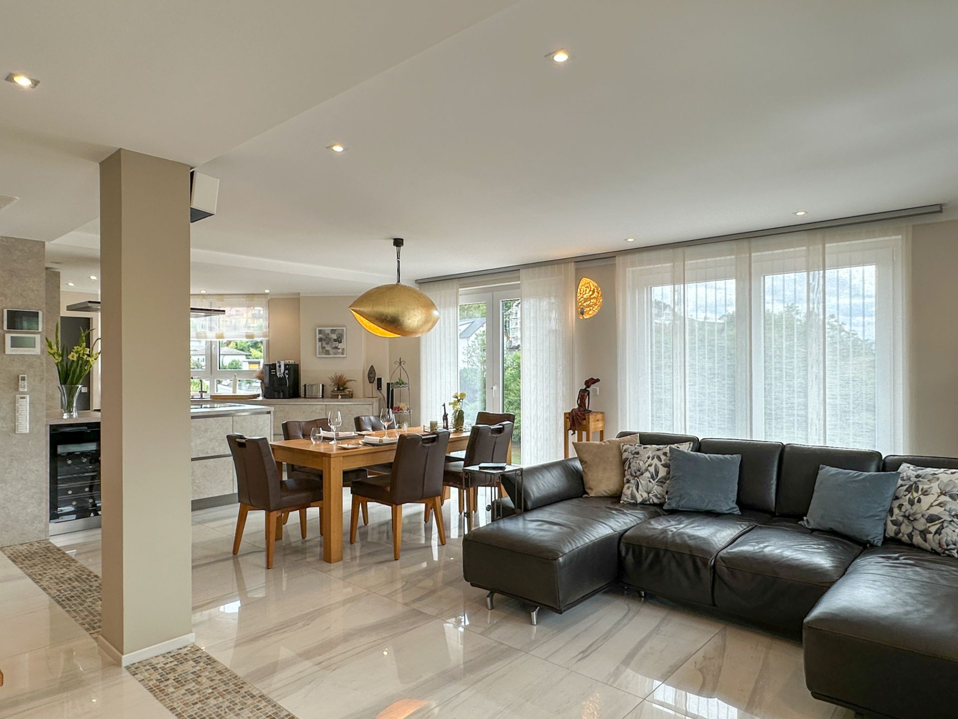 Helles Wohnzimmer mit schwarzem Sofa, Esstisch und großen Fenstern mit weißen Vorhängen.