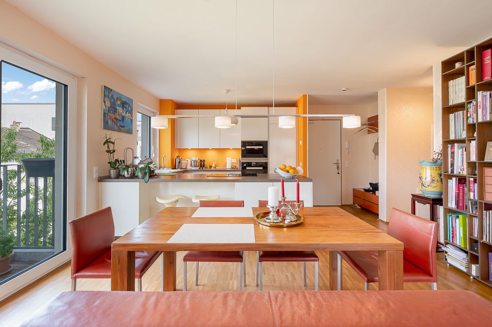 Helles Wohnzimmer mit Holztisch, rotem Stuhl, offener Küche und Bücherregal.