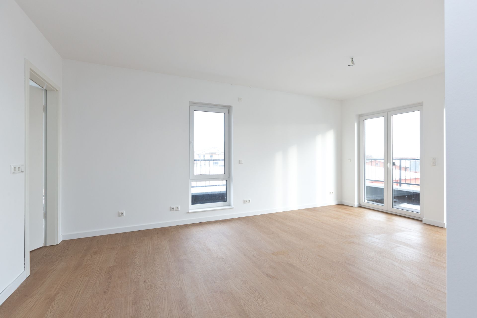 Heller, leerer Raum mit großem Fenster und Glastüren zu einem Balkon, Holzboden.