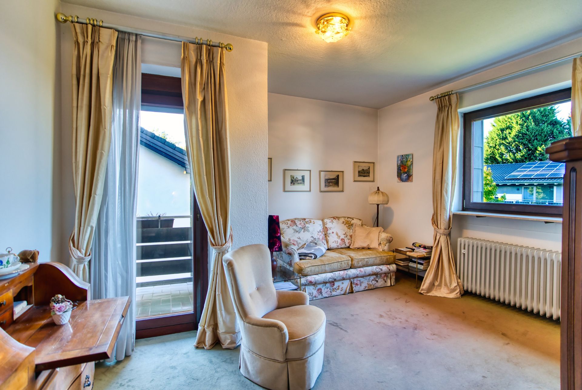 Wohnzimmer mit Sofa, Sessel, Schreibtisch, Vorhängen, Fenster und Tür zum Balkon.