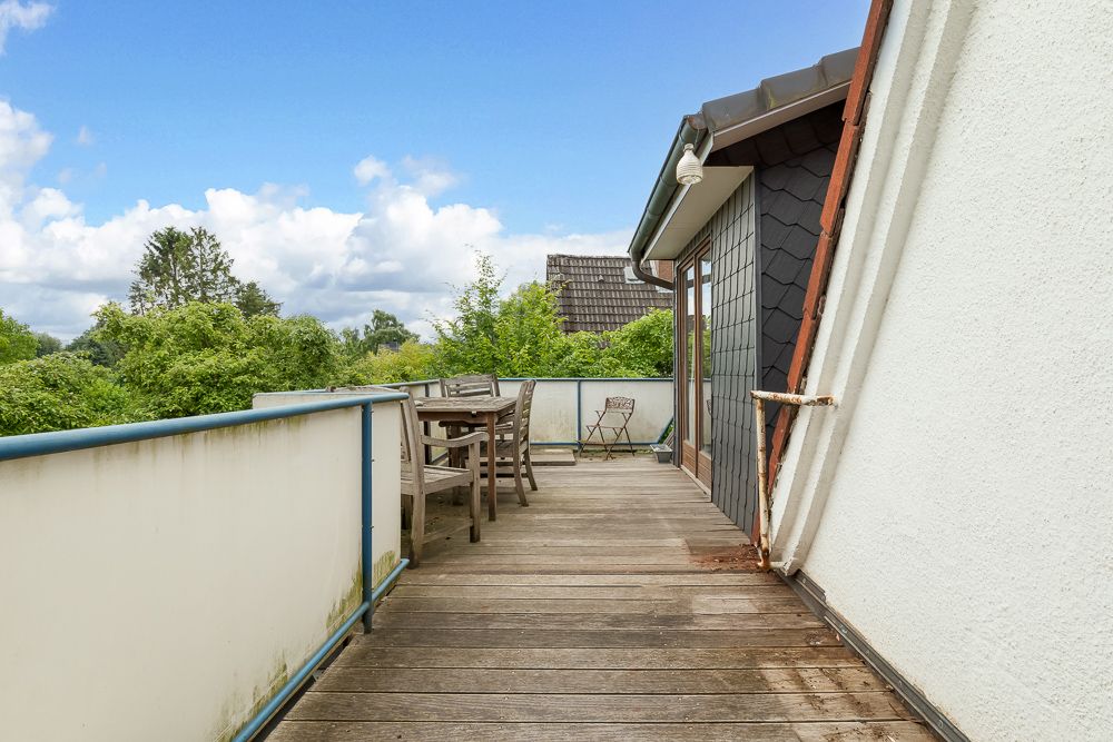 Holzterrasse mit Tisch und Stühlen, umgeben von grünen Bäumen unter blauem Himmel.
