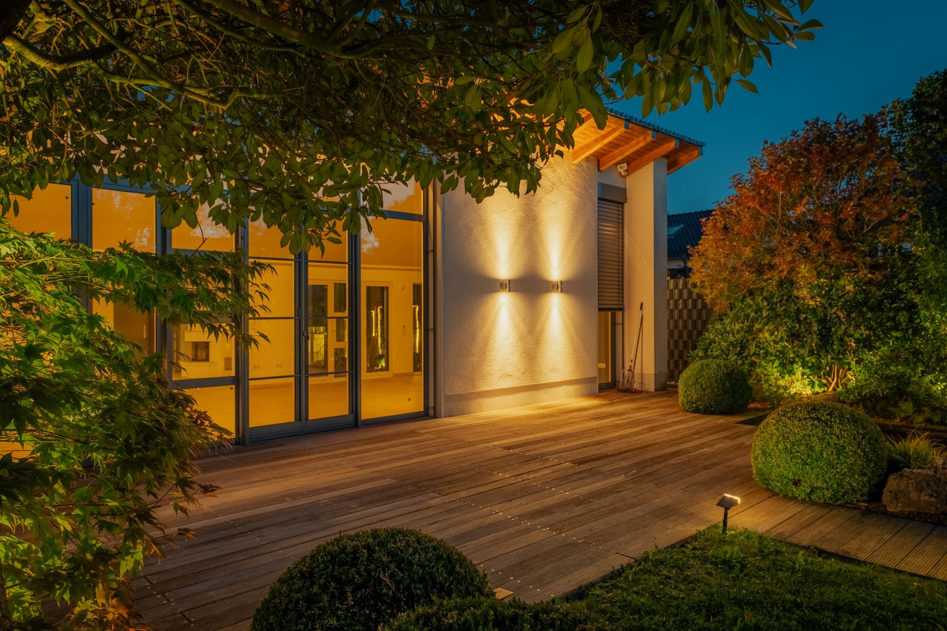 Beleuchtete Holzterrasse an einem modernen Wohnhaus mit großen Fenstern und Garten am Abend.