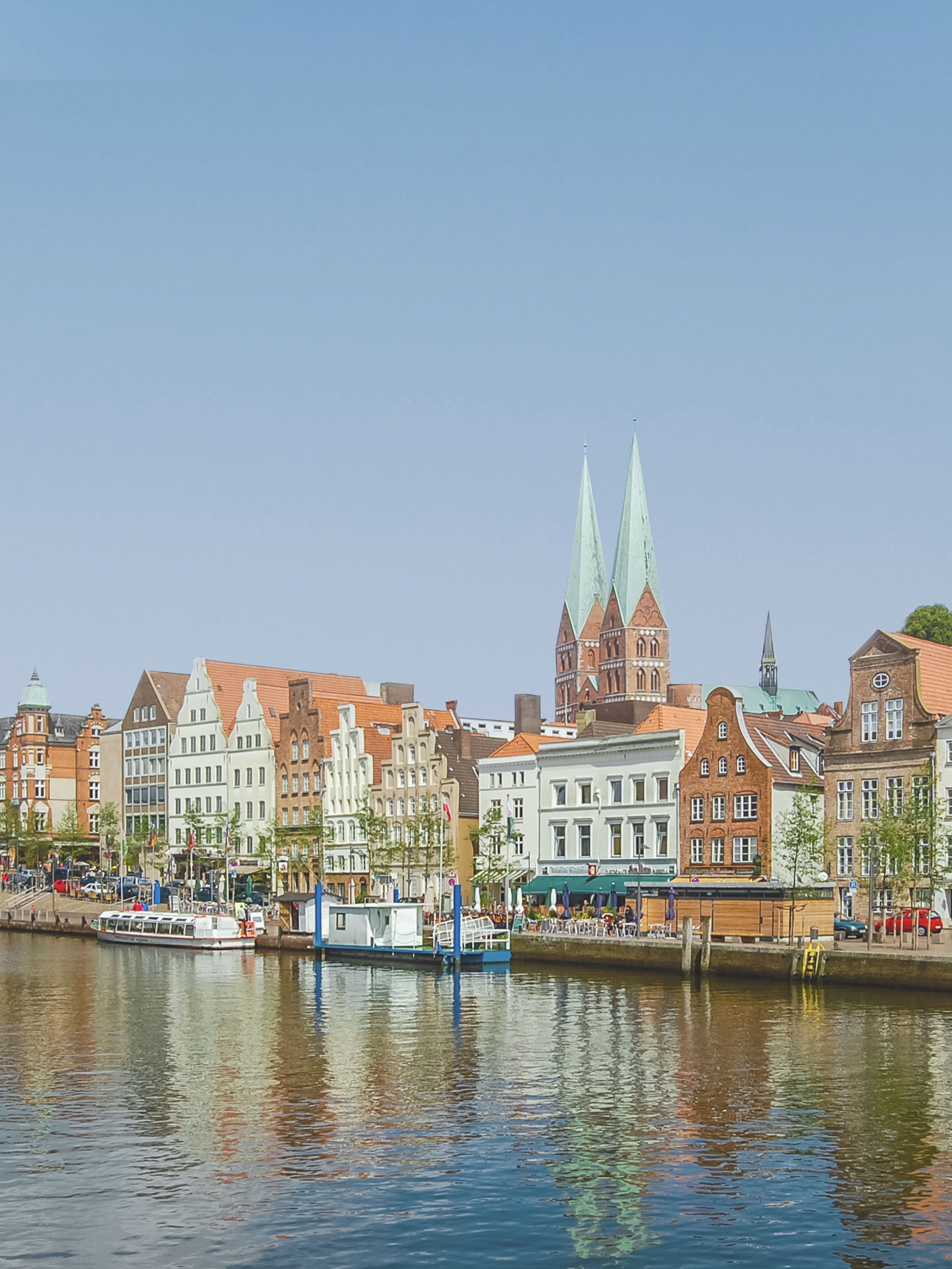 Altstadthäuser mit roten Dächern am Wasser, zwei Kirchtürme im Hintergrund.
