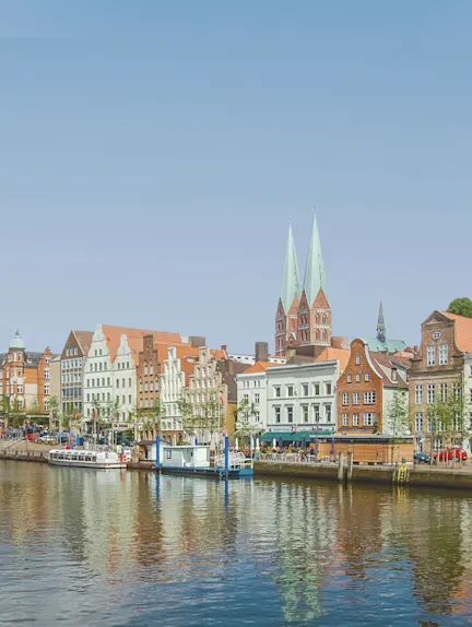 Altstadthäuser mit roten Dächern am Wasser, zwei Kirchtürme im Hintergrund.