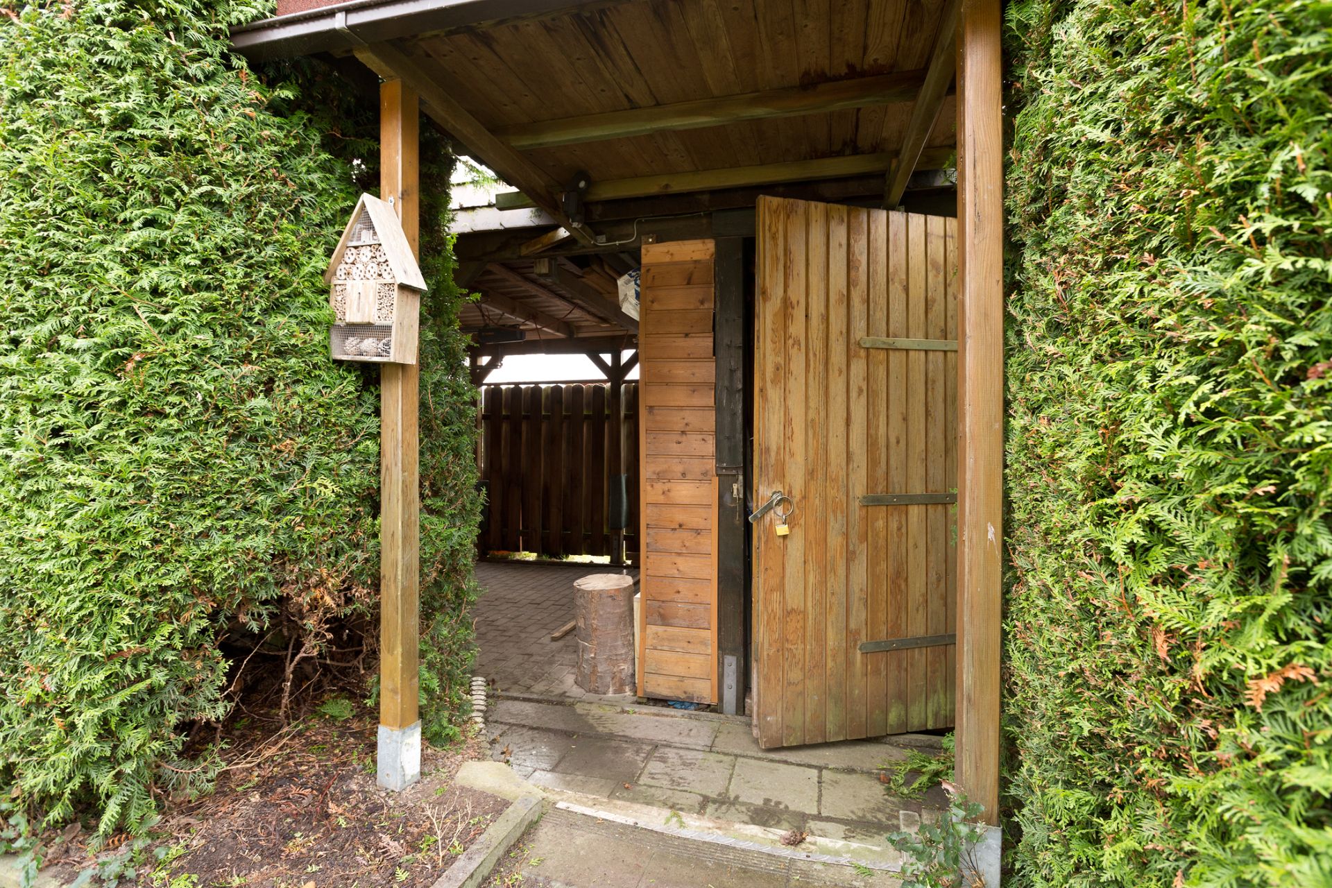 Holztür unter Überdachung im Garten zwischen hohen Hecken mit Vogelhaus an einem Pfosten.
