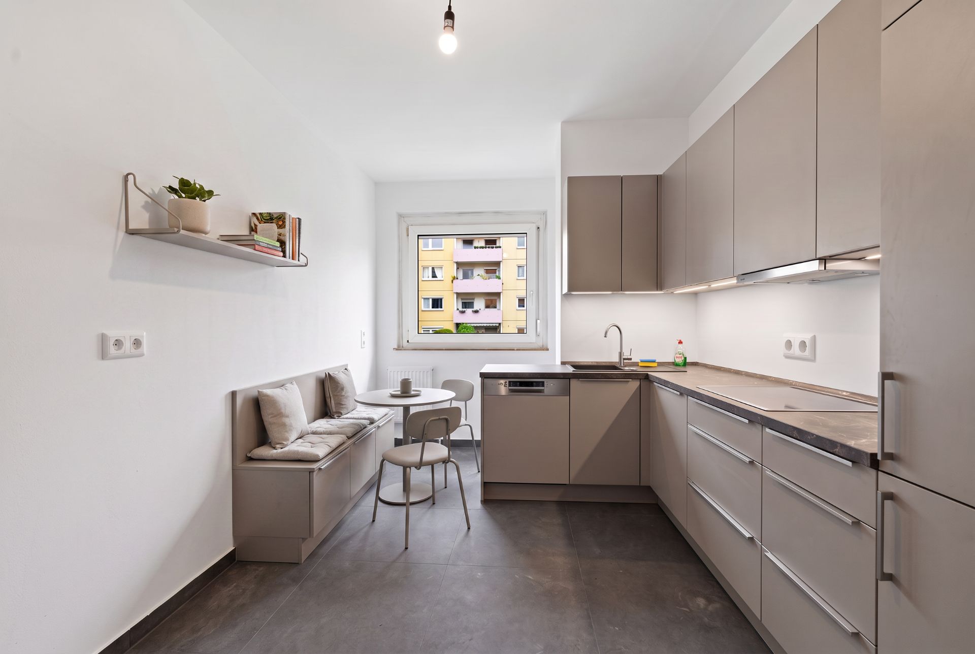 Moderne Küche mit grauen Schränken, Eckbank und kleinem Tisch an einem Fenster.