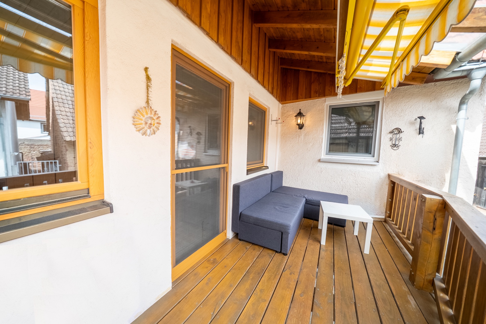 Überdachter Balkon mit Holzboden, Ecksofa, kleinem Tisch und Markise.