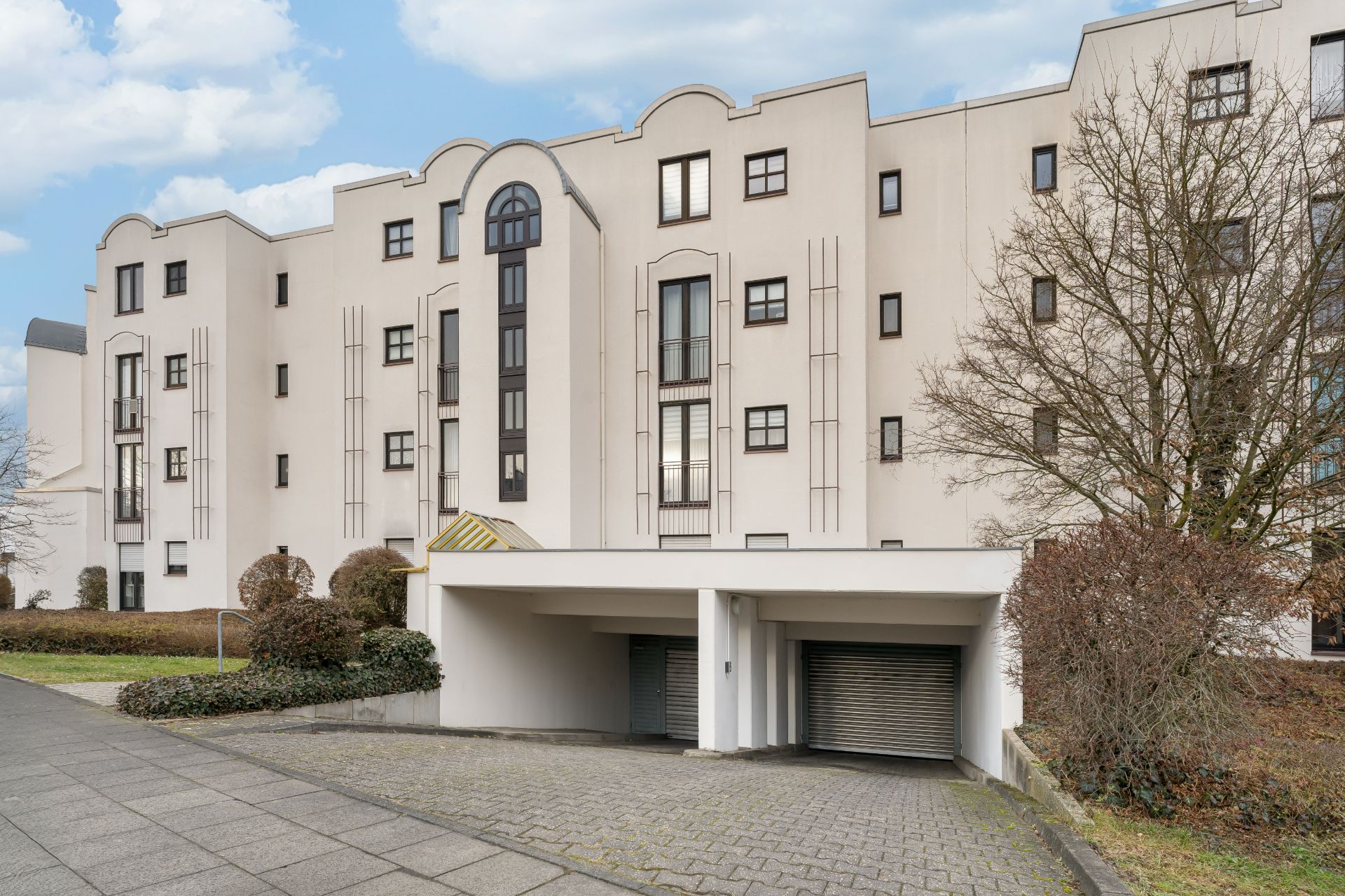 Mehrstöckiges Wohngebäude mit Garageneinfahrt und mehreren Fenstern, im Vordergrund ein Gehweg.