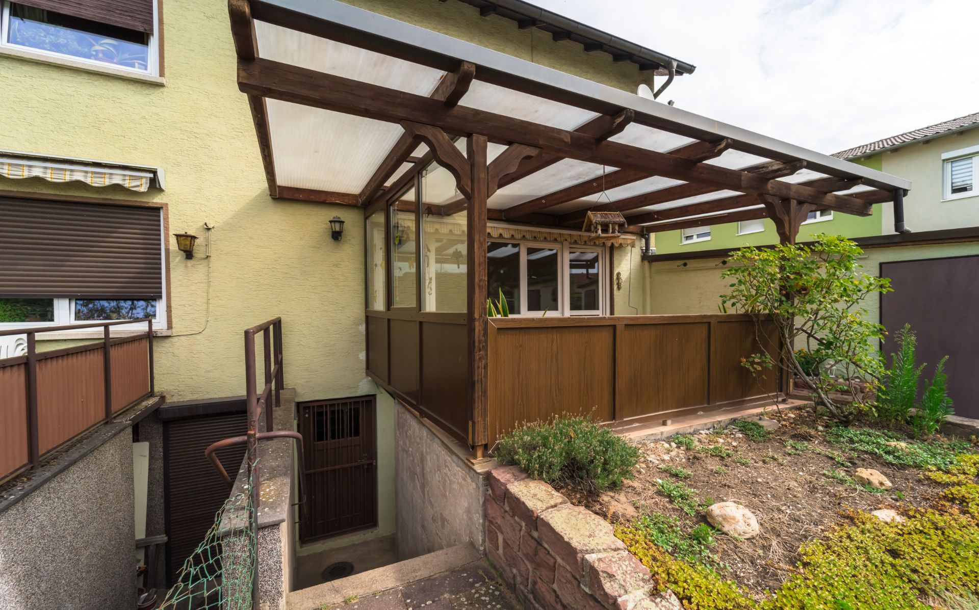 Überdachte Terrasse mit Holzverkleidung an einem Wohnhaus und kleiner Garten im Vordergrund.