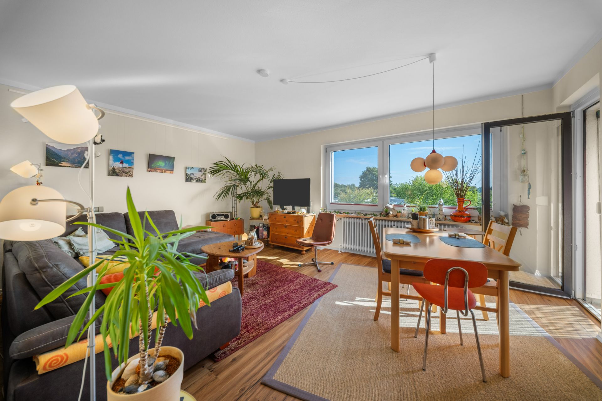 Helles Wohnzimmer mit Sofa, Esstisch, vielen Pflanzen und großen Fenstern mit Blick ins Grüne.