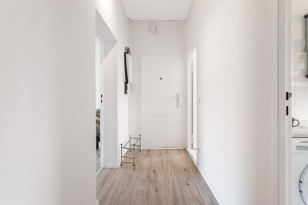 Heller Flur mit Holzboden, weißer Eingangstür und Schuhregal an der Wand.