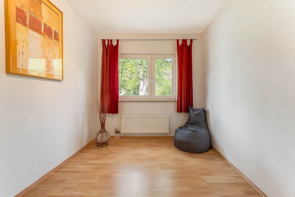 Kleiner, leerer Raum mit Holzfußboden, Fenster, rotem Vorhang, Sitzsack und Wandbild.
