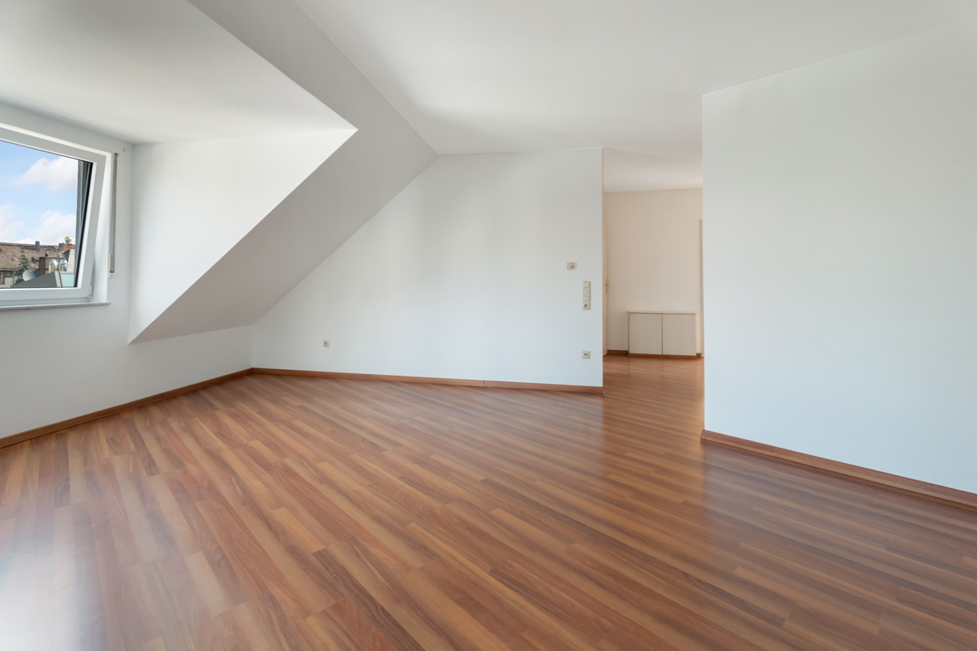 Helles, leeres Zimmer mit Holzfußboden und großem Fenster.