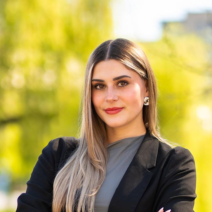 Junge Frau mit langen, blonden Haaren steht draußen vor grünen Bäumen.