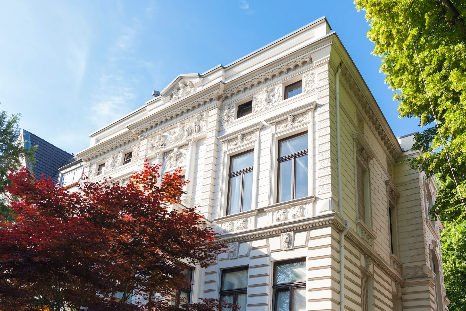 Weiße historische Villa mit Stuckfassade und großen Fenstern, Bäume im Vordergrund.