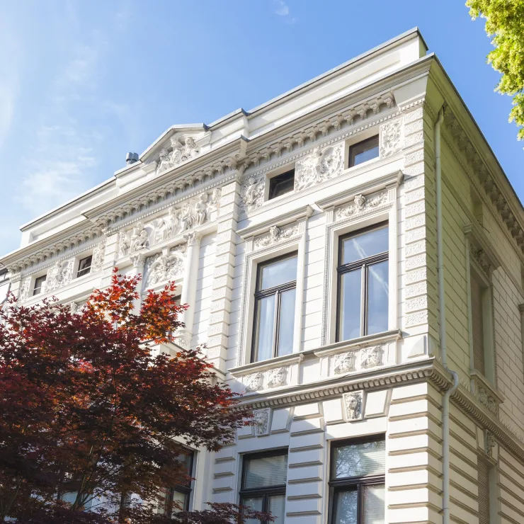 Weiße historische Villa mit Stuckfassade und großen Fenstern, Bäume im Vordergrund.