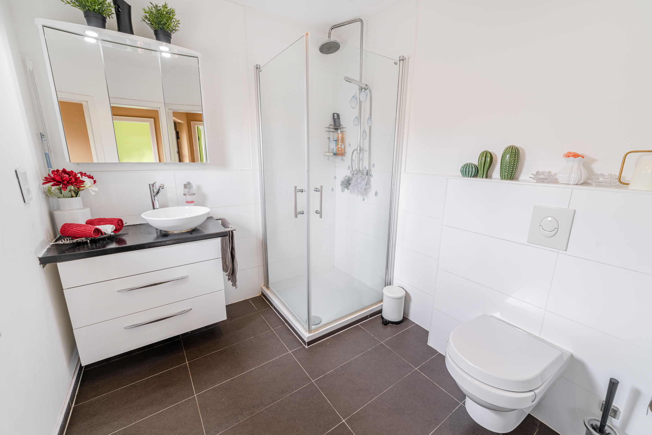 Modernes Badezimmer mit Glasdusche, Waschbecken, Wandtoilette und Dekoration auf weißen Fliesen.