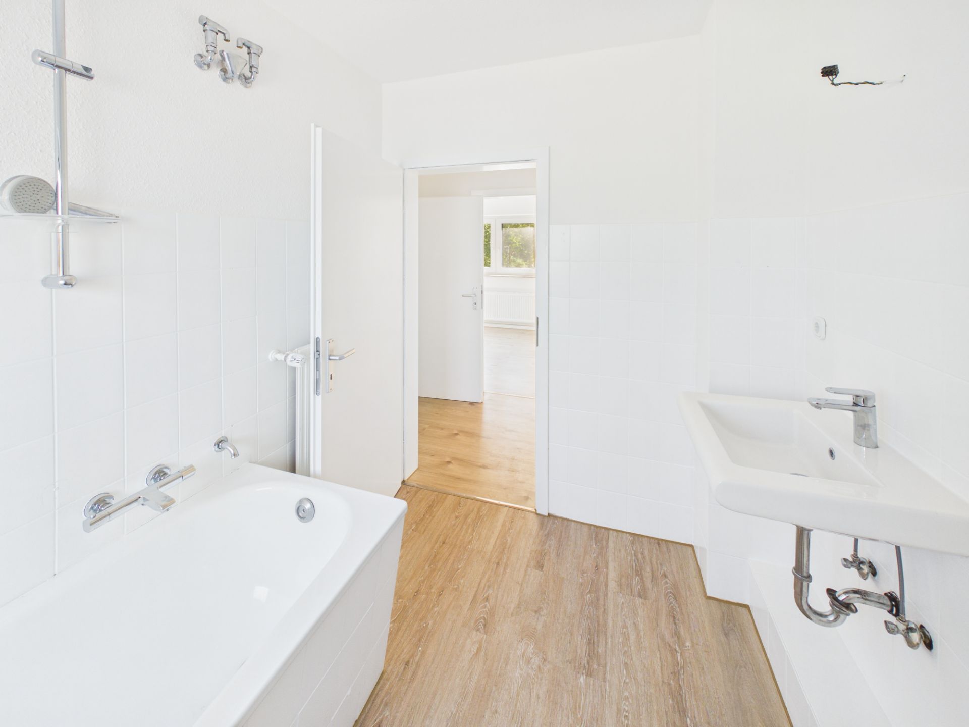Helles Badezimmer mit Badewanne, Waschbecken und Holzboden, Tür zum Flur offen.