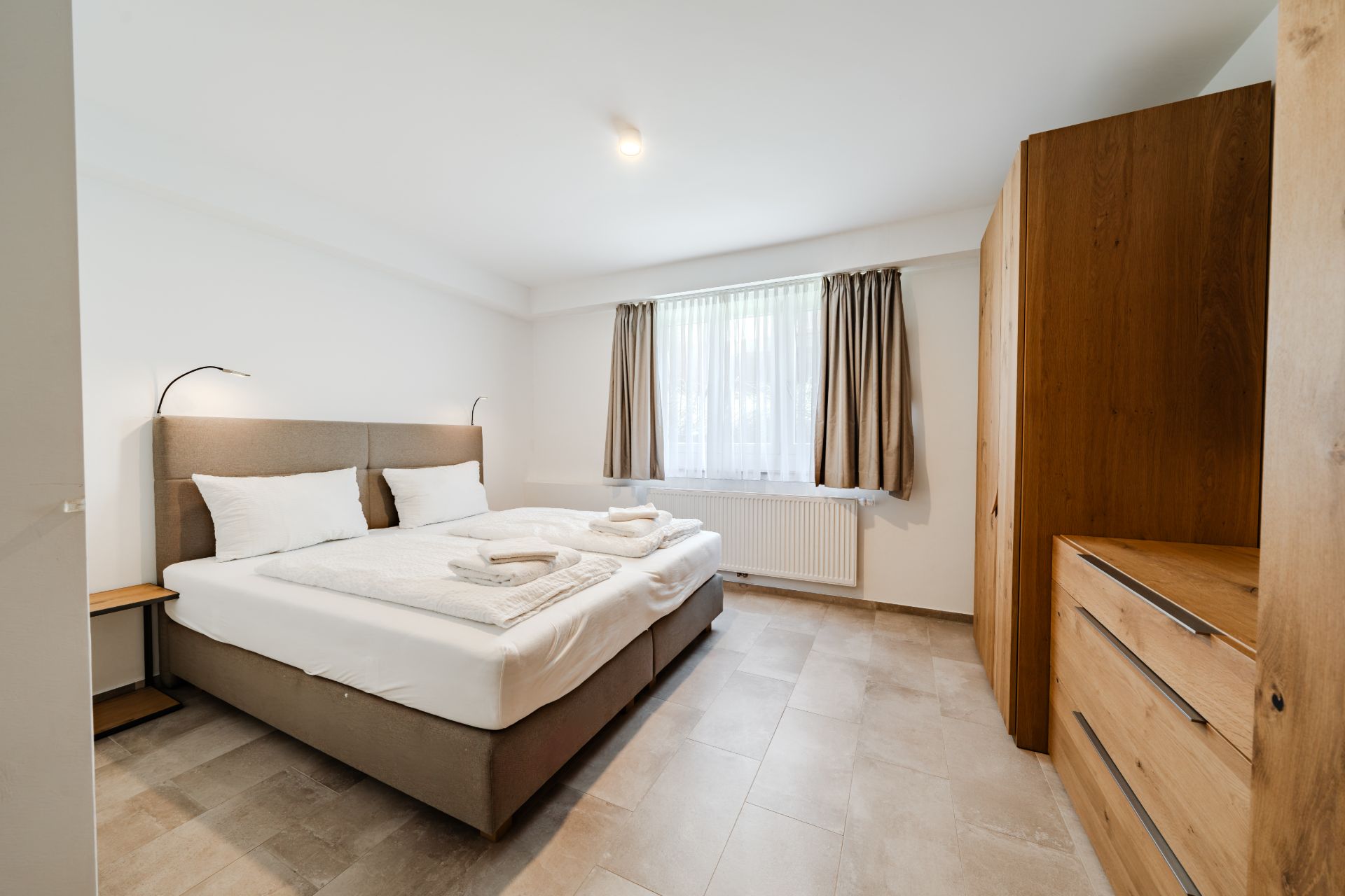 Schlafzimmer mit Doppelbett, Nachttischen, Holzschrank und Kommode, Fenster mit Vorhängen.