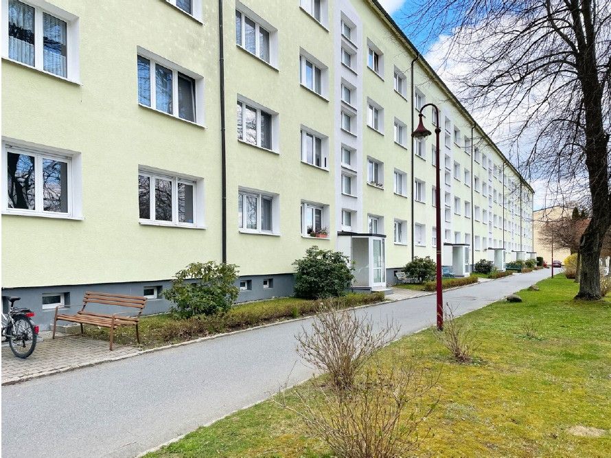 Mehrstöckiges Wohnhaus mit vielen Fenstern, Gehweg, Bank, Fahrrad und Vorgarten.