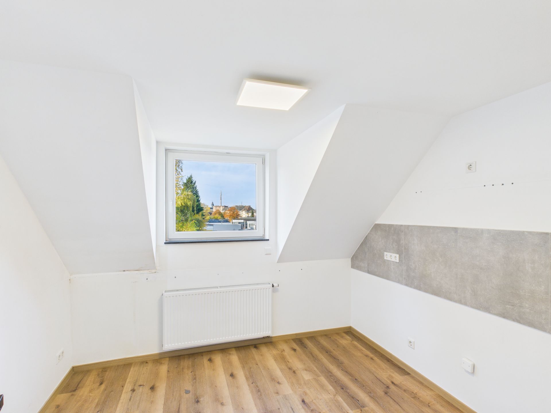 Helles Zimmer mit Holzfußboden, Dachschrägen und einem Fenster, darunter ein Heizkörper.
