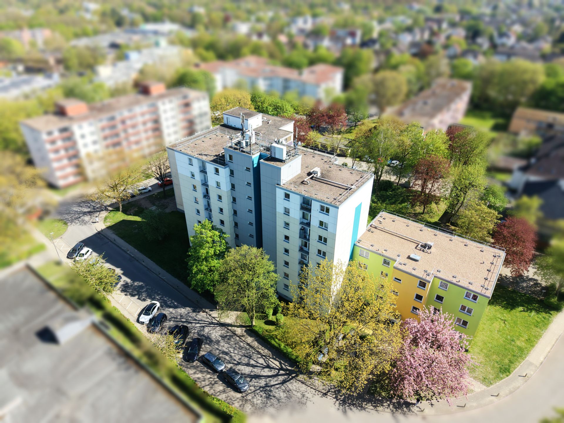 Mehrgeschossiges Wohnhaus umgeben von Bäumen und angrenzenden Straßen, daneben weiteres Haus.