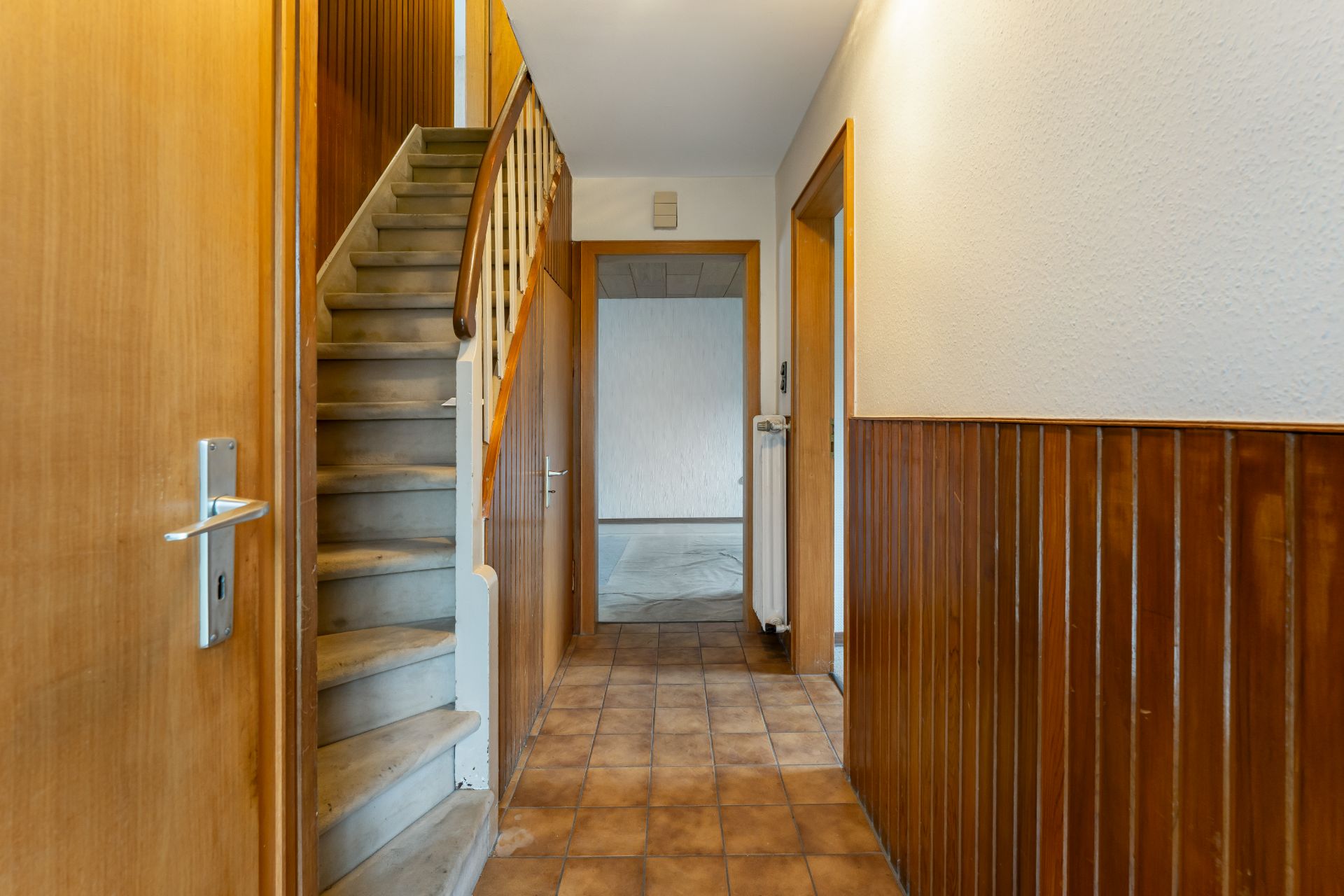 Hausflur mit braunen Fliesen, Holzvertäfelung, Treppe und offener Tür zu einem weiteren Raum.