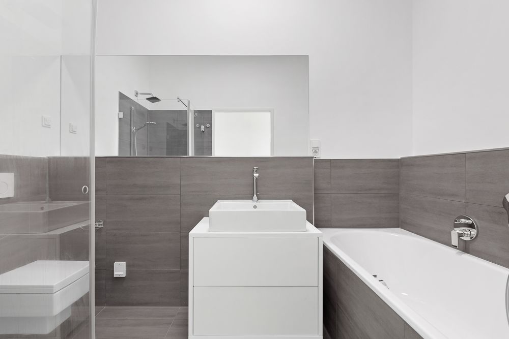 Modernes Badezimmer mit Badewanne, Waschbecken, Spiegel und Dusche, graue Fliesen und weiße Wände.