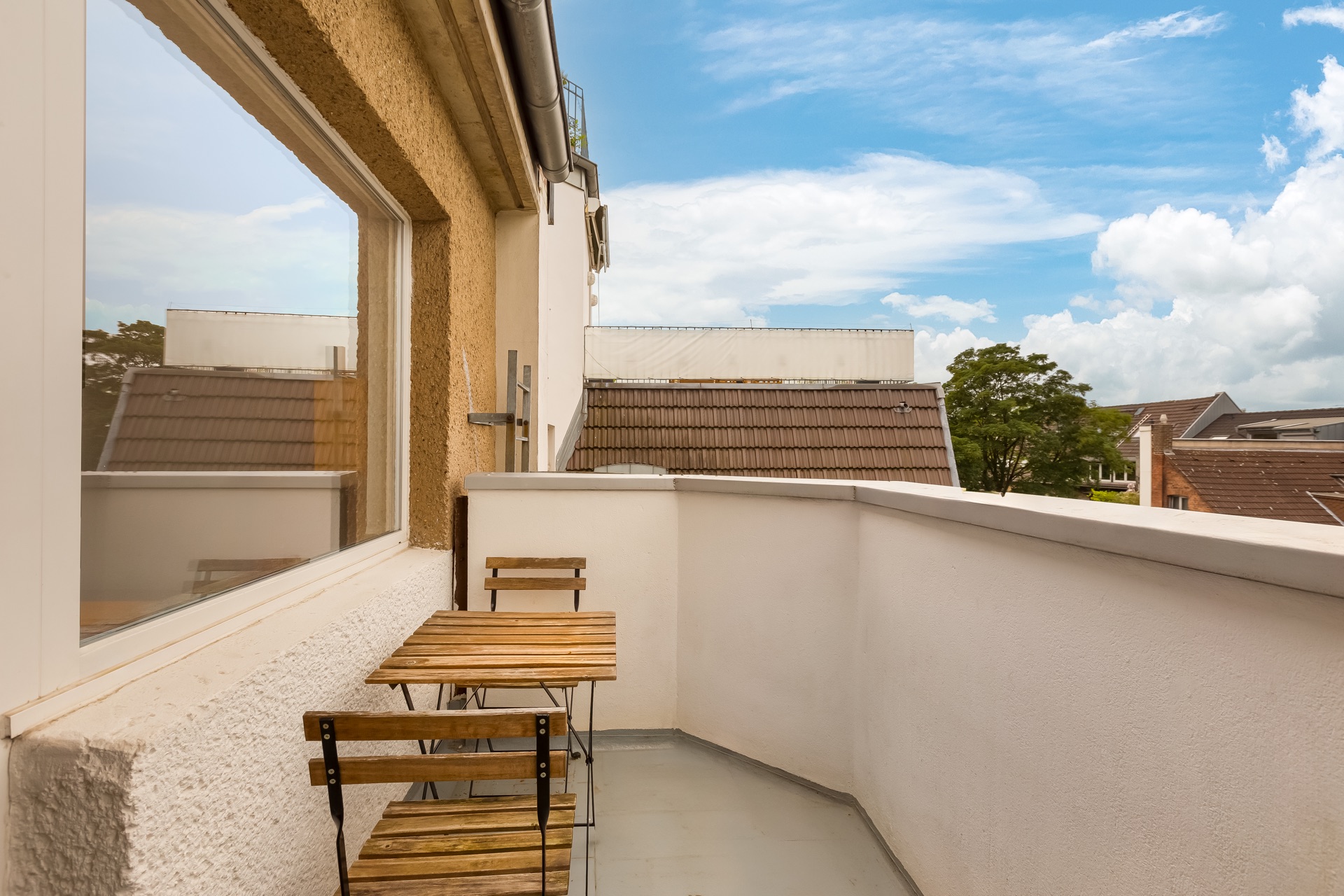 Kleiner Balkon mit Holztisch und zwei Stühlen, Blick auf Nachbarhäuser und blauen Himmel.