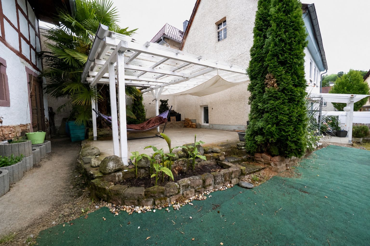 Überdachte Terrasse mit Hängematte, Hochbeet und Baum im Garten eines Wohnhauses.