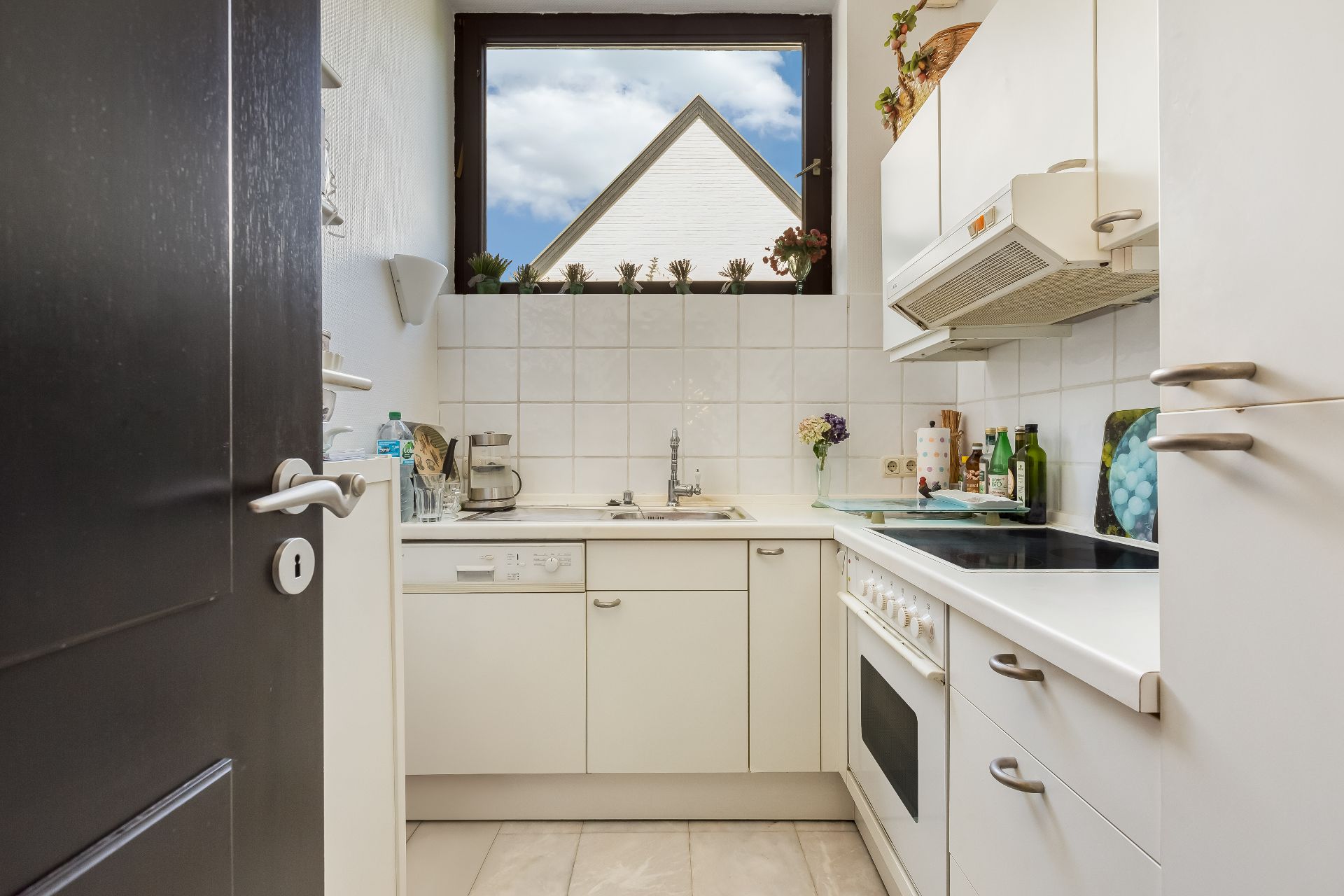 Helle, schmale Küche mit weißen Schränken, Kochfeld, Backofen und Fenster mit Blick nach draußen.