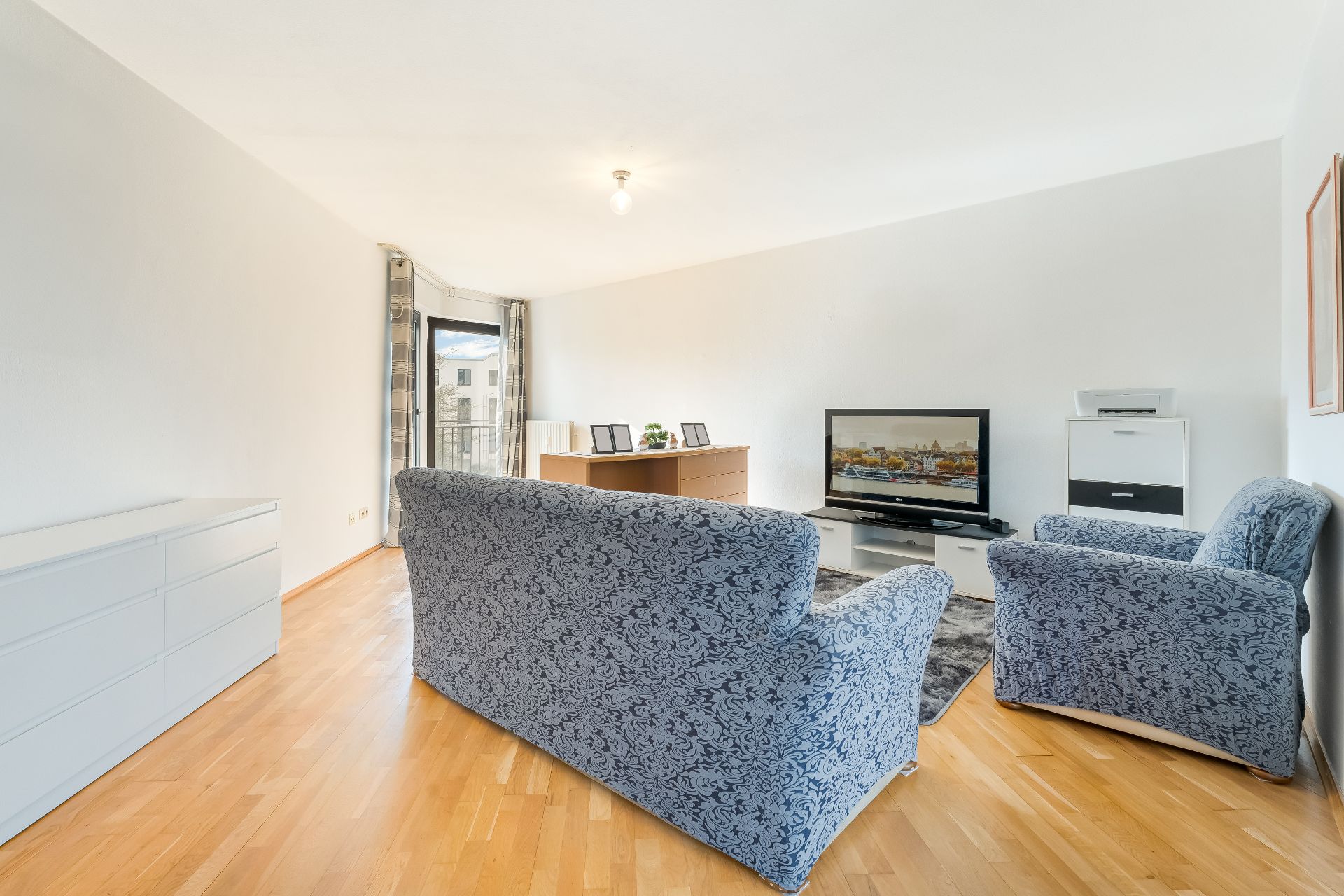 Wohnzimmer mit zwei blauen Sesseln, Fernseher, Kommode, Schreibtisch und Holzboden.