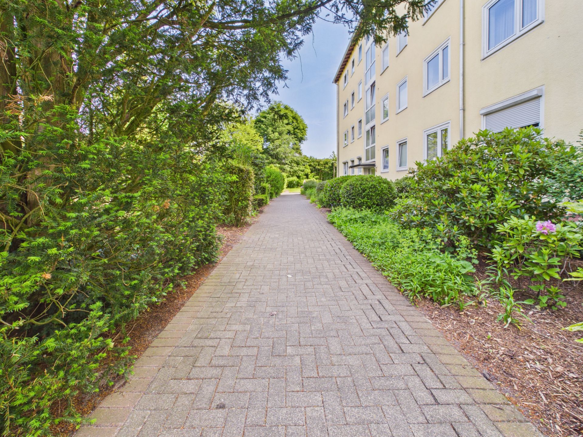 Gepflasterter Weg mit Büschen und Bäumen neben einem Mehrfamilienhaus.