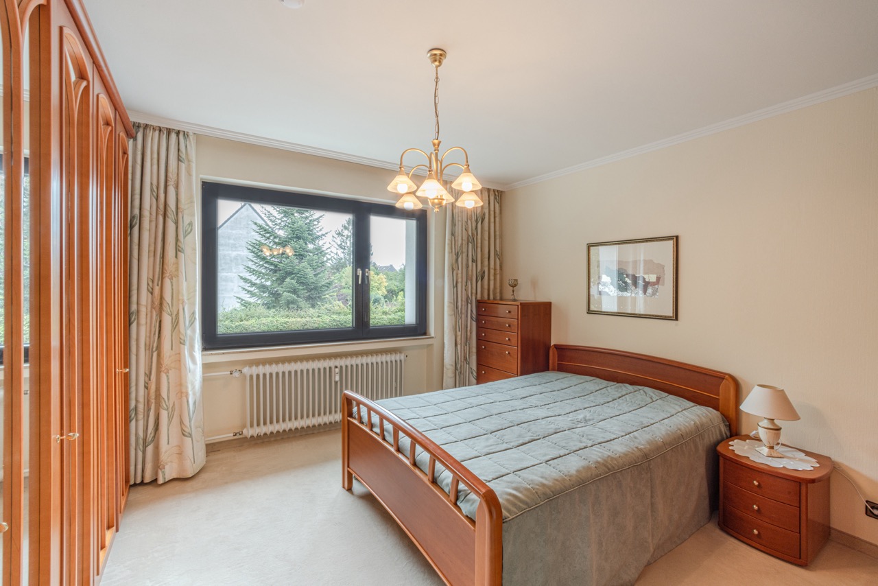 Helles Schlafzimmer mit Holzbett, Nachttisch, Kommode und großem Fenster mit Blick ins Grüne.