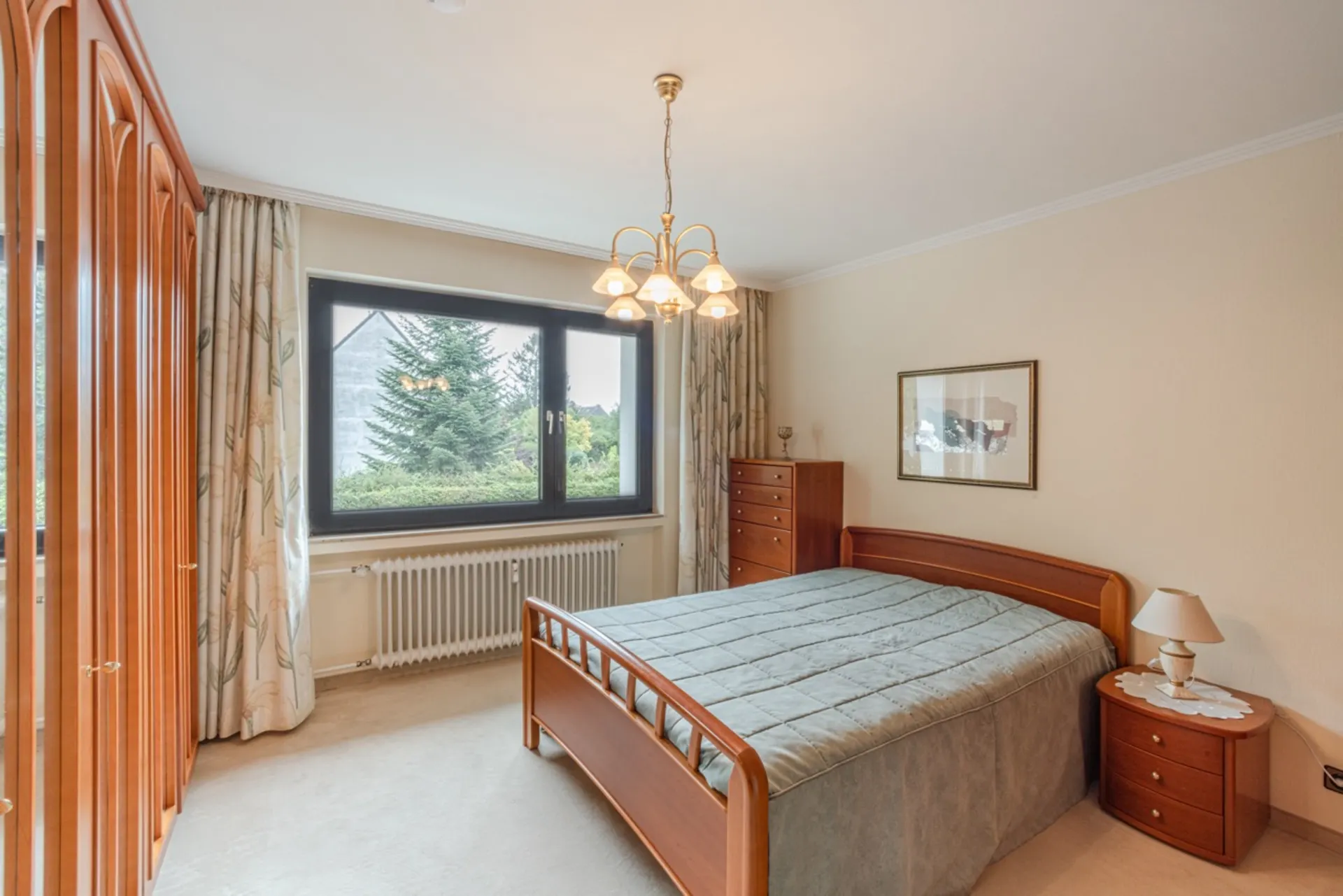 Helles Schlafzimmer mit Holzbett, Nachttisch, Kommode und großem Fenster mit Blick ins Grüne.